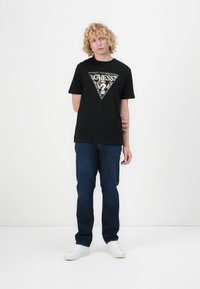 Svart bomull T-skjorte med en trekantet sølv- og gullpaljett «GUESS» logo, kombinert med mørkeblå jeans og hvite joggesko.