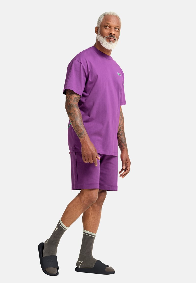 Homme âgé avec une barbe blanche portant un t-shirt violet et un short, des chaussettes grises et des sandales noires à glissière, debout sur un fond blanc.