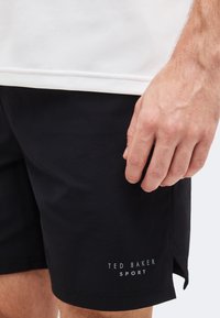 Κοντινή λήψη ενός χεριού που στηρίζεται κοντά στο κάτω μέρος μαύρων σορτς Ted Baker Sport, φορεμένα με λευκό πουκάμισο.