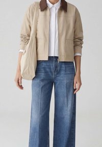 Persoon draagt een beige jas met een bruine kraag, een wit overhemd, blauwe wijde jeans en draagt een beige schoudertas.