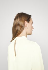Blusa de manga longa branca com um detalhe em buraco na parte de trás, estilizada com brincos de argola transparentes; cabelo solto sobre um dos ombros.