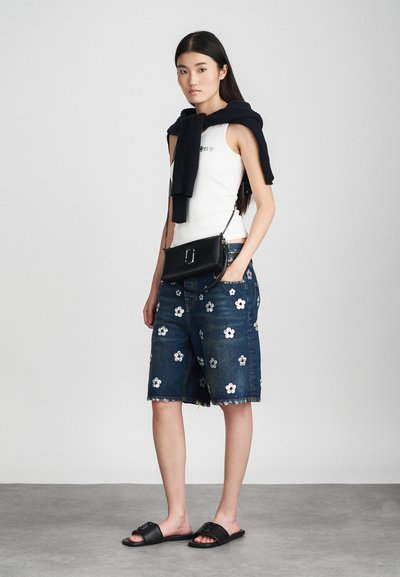 Bermuda shorts in denim con ricami floreali, canotta bianca, maglione nero di lana appoggiato sulle spalle e sandali neri a scivolo.