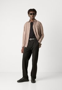 Camicia button-up in lino marrone chiaro sopra una maglietta nera, pantaloni neri a gamba larga e sneakers nere, accessoriate con occhiali da sole scuri.