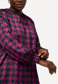 Langærmet bluse i lilla med et houndstooth-mønster i mørkegrøn. Har en samlet halsudskæring og smockede manchetter for en afslappet pasform.