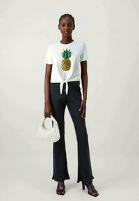T-shirt branca com uma estampa de ananás e nó à frente, combinado com calças pretas largas. Acessórios incluem uma mala branca e sandálias azuis.