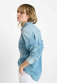 Lichtblauw denim overhemd met lange mouwen, met knoopsluiting bij de manchetten en een borstzak. Heeft een afgeronde zoom en een klassiek kraagontwerp.