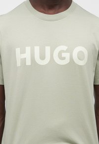 Άνδρας που φοράει ανοιχτοπράσινη μπλούζα με μεγάλο λευκό κείμενο "HUGO" στο στήθος.