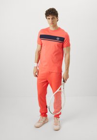 Tenisový outfit se světlou korálovou košilí s krátkým rukávem a námořnickým pruhem, odpovídajícími korálovými kalhotami a světlými teniskami, držící tenisovou raketu.
