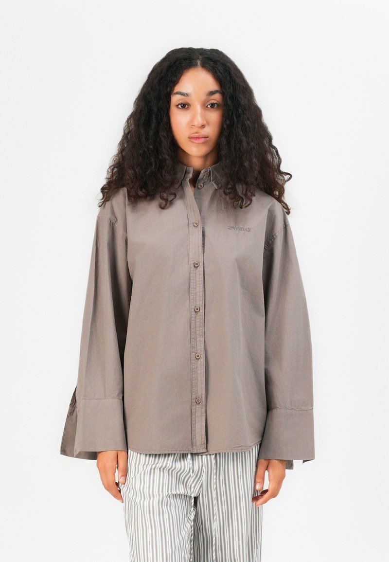 Brown long-sleeve shirt with a button-down front, straight cut, and a relaxed fit. Features a subtle logo on the chest and flared cuffs.