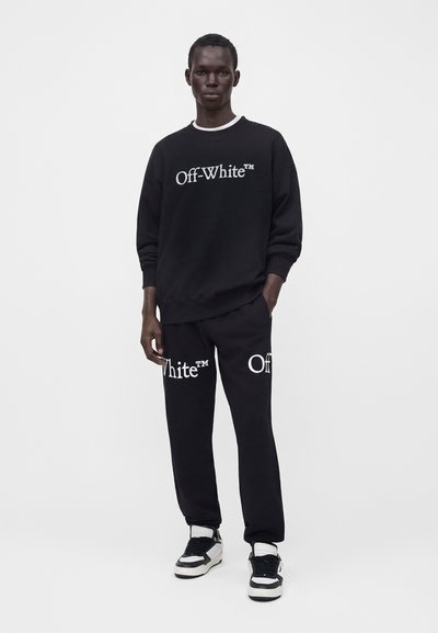 Άνδρας που φοράει μαύρο φούτερ Off-White και ταιριαστό παντελόνι με λευκό λογότυπο, στέκεται μπροστά σε μονόχρωμο λευκό φόντο.