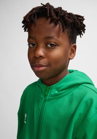 Grøn zip-up hoodie lavet af blødt stof, med en høj krave og et lille hvidt logo på venstre side af brystet.