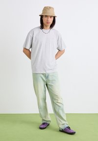 Grå oversized t-skjorte, lys blå-grønne slitte jeans, lilla sko, beige bøttehatt og fargerik perlekjede. Enkelt bakgrunn.