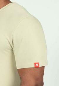 Beige katoenen t-shirt met korte mouwen en ronde hals, met een klein rood etiket met een wit leeuwenlogo nabij de zoom. Gladde textuur.