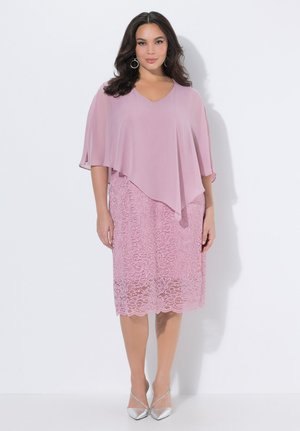 Frau trägt ein knielanges pinkfarbenes Spitzenkleid mit asymmetrischem Überwurf und silbernen spitzen Absätzen, steht vor einem schlichten weißen Hintergrund.