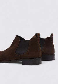 Bottines en suède marron avec des inserts élastiques sur les côtés, bout arrondi et petit talon. Équipées de tirettes à l'arrière pour un enfilage facile.
