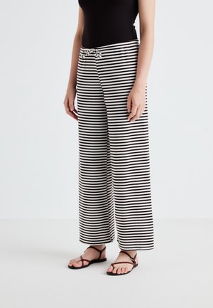 Pantalon à rayures noires et blanches, large et à taille élastique avec un détail en nœud à l'avant, associé à des sandales plates.
