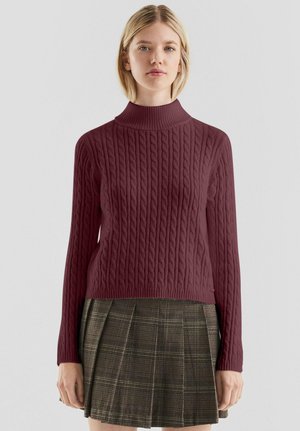 Woman wearing a burgundy cable-knit turtleneck sweater and a brown plaid pleated skirt, standing against a plain background.