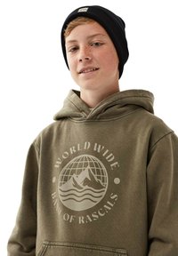 Olivgrön hoodie med ett stort vitt grafiskt mönster som föreställer berg och vågor. Åtföljd av en svart stickad mössa.
