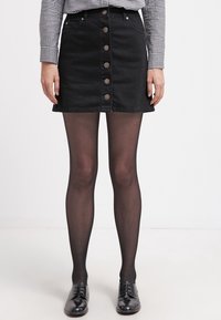 Svart denim A-linje kjol med framsida knappar, kombinerad med svarta ogenomskinliga strumpbyxor och glänsande svarta skor, mot en vit bakgrund.