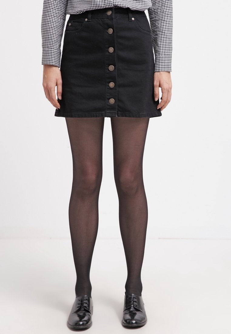 Svart denim A-linje kjol med framsida knappar, kombinerad med svarta ogenomskinliga strumpbyxor och glänsande svarta skor, mot en vit bakgrund.