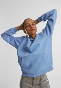 Svetlo modrá mikina s uvoľneným strihom, kapucňou a prednou vreckom. Vyrobená z mäkkej tkaniny, s rebrovanými manžetami a dolným okrajom, s hladkou textúrou.