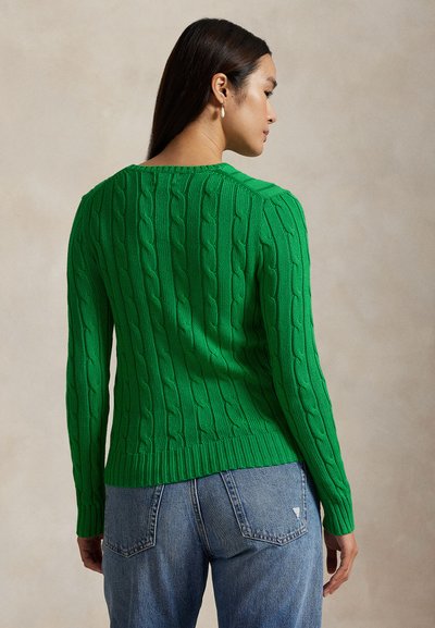 Femeie cu păr lung și închis la culoare, purtând un pulover verde cu model împletit și blugi albaștri, surprinsă din spate pe un fundal simplu.