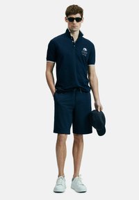 Man in een marineblauwe polo met bijpassende korte broek, witte sneakers, zwarte zonnebril en houdt een marineblauwe pet in één hand.