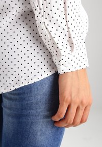 Camicia bianca con pois neri, dotata di polsino con bottoni e dettagli con maniche plissettate, indossata sopra jeans in denim blu.