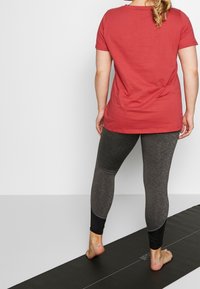 Camiseta de algodón roja de manga corta combinada con leggings texturizados de color gris oscuro con acentos en negro. Descalzo sobre una esterilla de yoga.