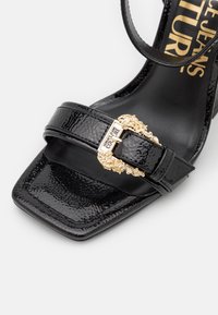 Sandale en cuir verni noir à bout carré, boucle dorée ornée et marquage "Versace Jeans Couture" sur la semelle intérieure et la boucle.