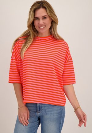 Blonde vrouw die glimlacht, draagt een los rood en roze gestreept T-shirt en lichtblauwe spijkerbroek, tegen een effen beige achtergrond.