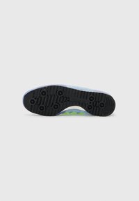 Suela de zapatilla con goma negra, seis tacos de tracción. Parte superior en color azul claro con acentos verdes, superficie texturizada y bordes suaves.