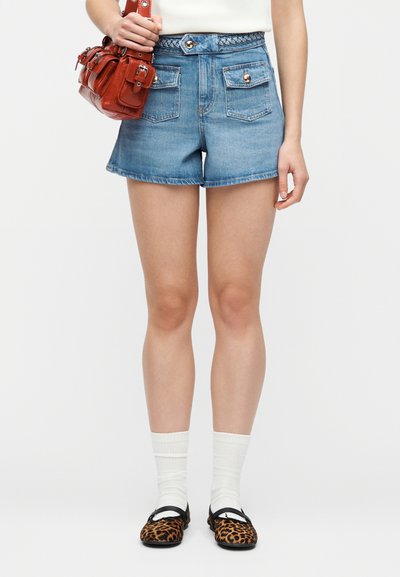Γυναίκα που φοράει μπλε τζιν σορτς ψηλόμεσα με τσεπάκια με κουμπιά, λευκές κάλτσες, μπαλαρίνες με λεοπάρ σχέδιο και κρατά μια μικρή καφέ δερμάτινη τσάντα.