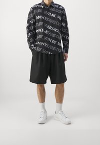 Svart skjorta med knappar och ett mönster av fet vit text, i kombination med lösa svarta shorts. Avslutas med vita sneakers och strumpor som går upp till vristerna.