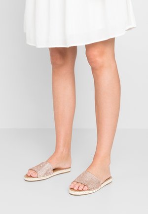 Kvinde iført hvid kjole og glitrende beige slip-on sandaler stående på gråt gulv.