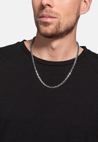 Collana di catena d'argento con anelli testurizzati, indossata su una camicia nera, caratterizzata da un design piatto e schemi alternati nel metallo.
