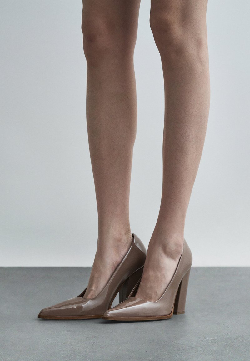 Tacones altos de charol beige con punta afilada y un diseño recortado, que presentan un grueso tacón en bloque y un acabado liso y brillante.
