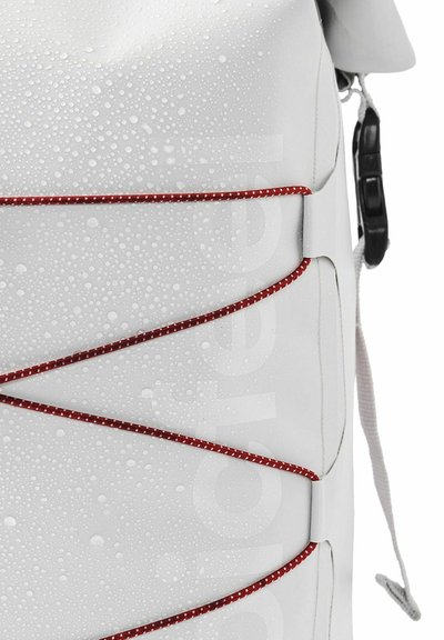 Sac imperméable blanc avec des accents élastiques rouges, présentant une surface texturée avec des gouttes d'eau. Matériel réglable sur le côté.