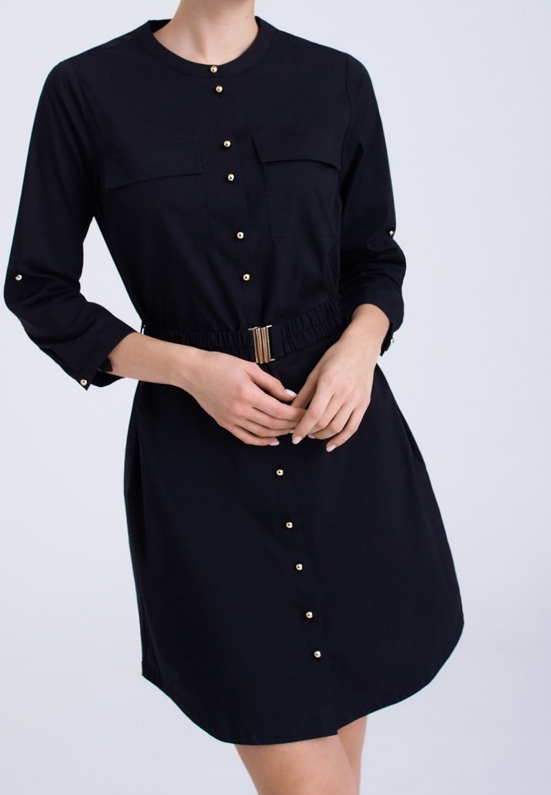 Svart klänning med långa ärmar, knäppning fram, två bröstfickor, elastisk midja och guldknappdetaljer. Slät tyg, knä-lång.