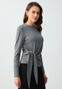 Femme aux cheveux châtain moyen portant un haut gris à manches longues noué devant et une jupe noire, debout sur un fond uni.