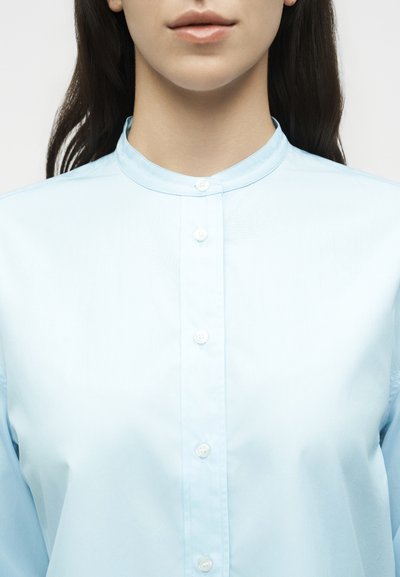 Γυναίκα με ανοιχτό μπλε πουκάμισο με μανταρίνι γιακά και σκούρα μαλλιά σε λευκό φόντο.