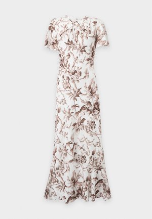 Kvinde iført en hvid maxikjole med brune blomsterprint, korte ærmer og beige højhælede sandaler, der står mod en ensfarvet baggrund.