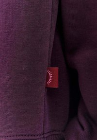 Fialová bavlněná mikina s texturovanou tkaninou, na které se nachází červená nášivka s vyšívaným růžovým detailem. Viditelné jsou čisté linie a jemné švy.