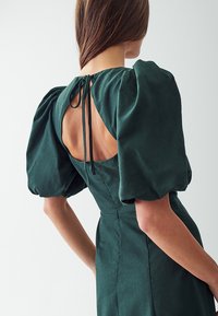 Vihreä, puffihihainen mekko, jossa on avoin selkäosa ja kaulassa solmittava nauha. Se on valmistettu teksturoidusta kankaasta, ja siinä on muotoiltu siluetti sekä saumat. 