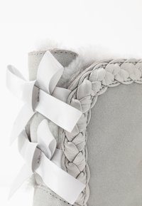 Szare zamszowe botki z plecionymi detalami wzdłuż krawędzi, z dwoma białymi satynowymi kokardkami po bokach i miękką, puszystą podszewką wewnętrzną.