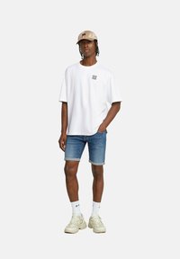 T-shirt bianca in cotone con una grafica quadrata sul lato sinistro del petto, abbinata a pantaloni corti in denim blu. Un cappellino beige e sneakers beige chiaro completano il look.