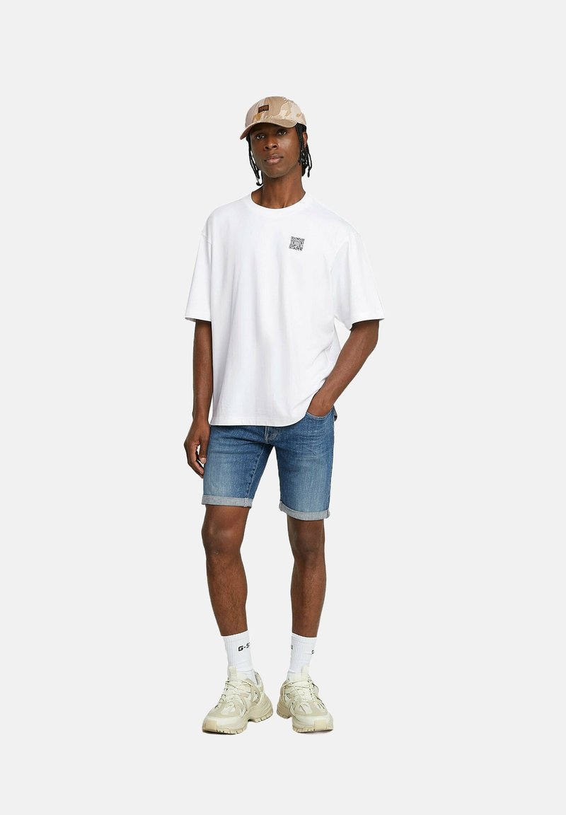 T-shirt bianca in cotone con una grafica quadrata sul lato sinistro del petto, abbinata a pantaloni corti in denim blu. Un cappellino beige e sneakers beige chiaro completano il look.