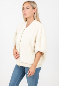 Veste en polaire couleur crème avec une fermeture éclair sur le devant, des manches surdimensionnées et un ourlet côtelé. Comprend des poches latérales et une finition en tissu texturé.
