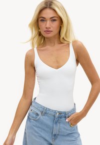 Femme blonde portant des boucles d'oreilles en or, un top blanc sans manches à col en V et un jean ample bleu clair, la main droite dans la poche.