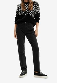 Schwarzer Pullover mit weißem Zebramuster, kombiniert mit hochgeschnittenen schwarzen Jeans mit Perlenakzenten und schwarzen Sneakers mit weißen Sohlen.